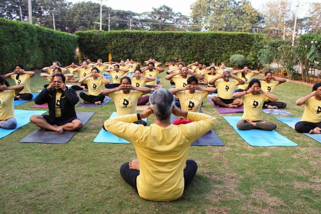 a group of students practicing pranayama in a open garden as a part of a course in a yoga school led by a teacher