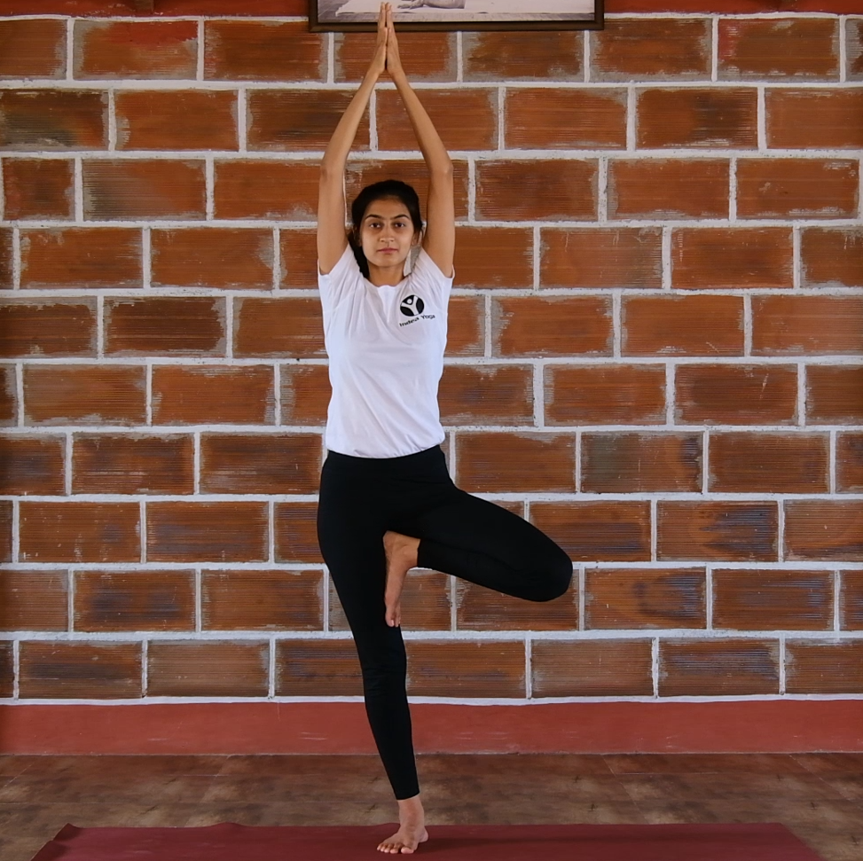 Vrkshasana (Tree Posture) involves balancing on one leg performed by a girl in white tshirt and black pants with a brick wall backdrop.