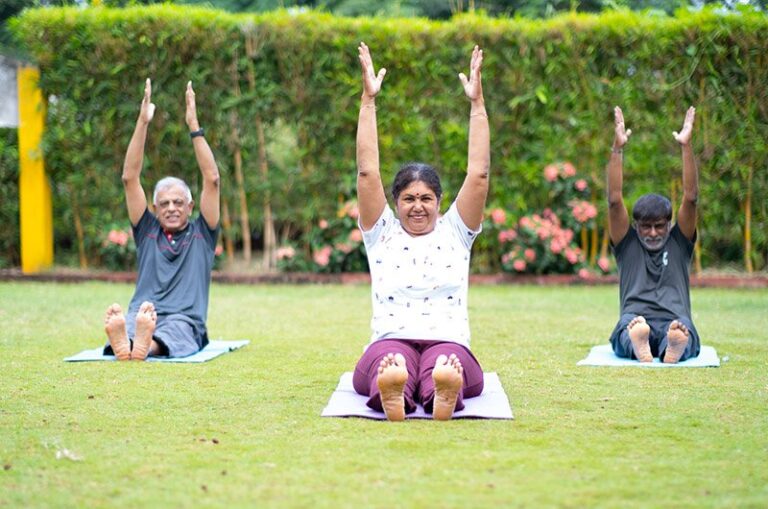 Three elderly people practicing therapy yoga in an outdoor garden, smiling and enjoying a peaceful wellness session at Arogyam.