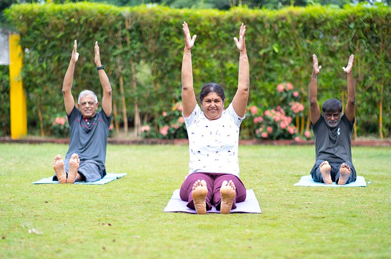 Three elderly people practicing therapy yoga in an outdoor garden, smiling and enjoying a peaceful wellness session at Arogyam.