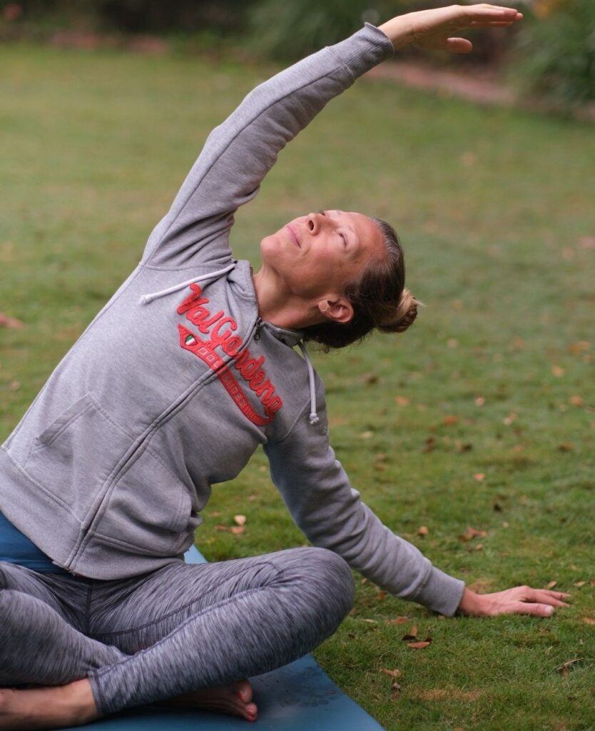 a woman sitting in a garden and bending to her left with arms extended out. with a pleasant smile on her face.