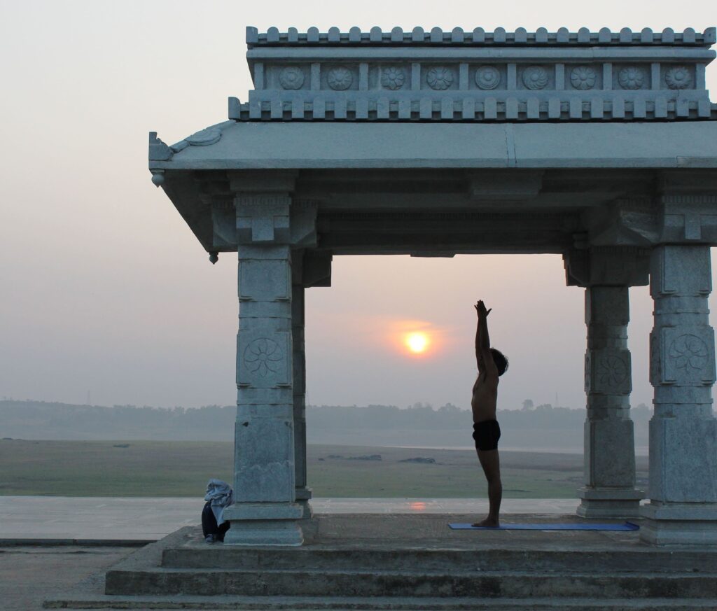 bharat shetty in a beautiful Indian temple mandap close to a river practicing sun salutation.